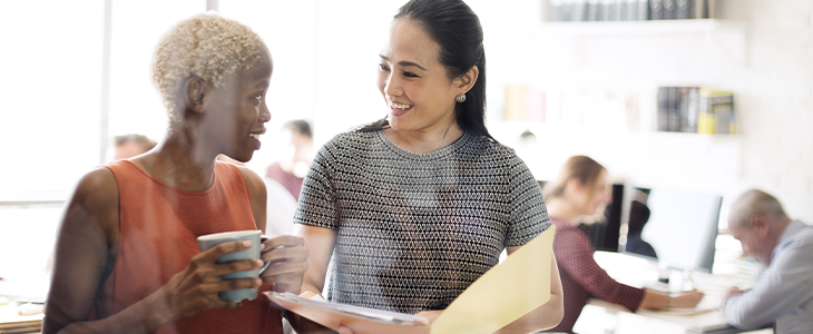 Two women talking. One is holding a cup of coffee and the other is holding a file open.