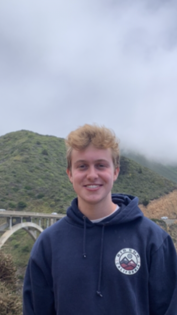 A person smiling with a mountainous landscape and bridge in the background.