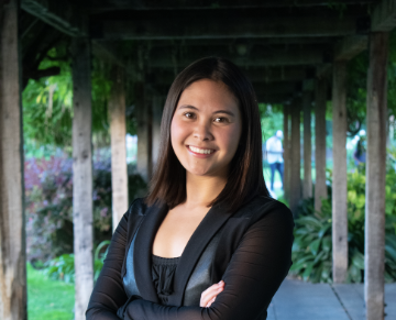 A person smiling with arms crossed, standing under a wooden pergola.