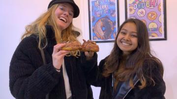 Two people smiling and holding up peace signs in front of framed pictures.