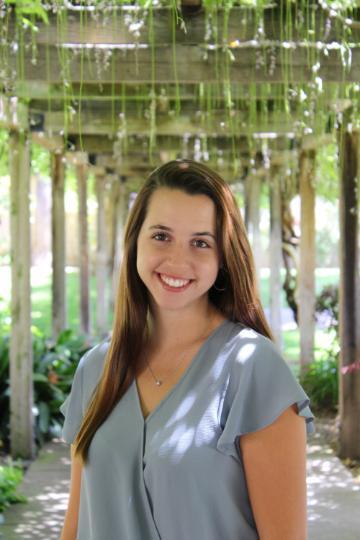 A person smiling outdoors with a greenery background.