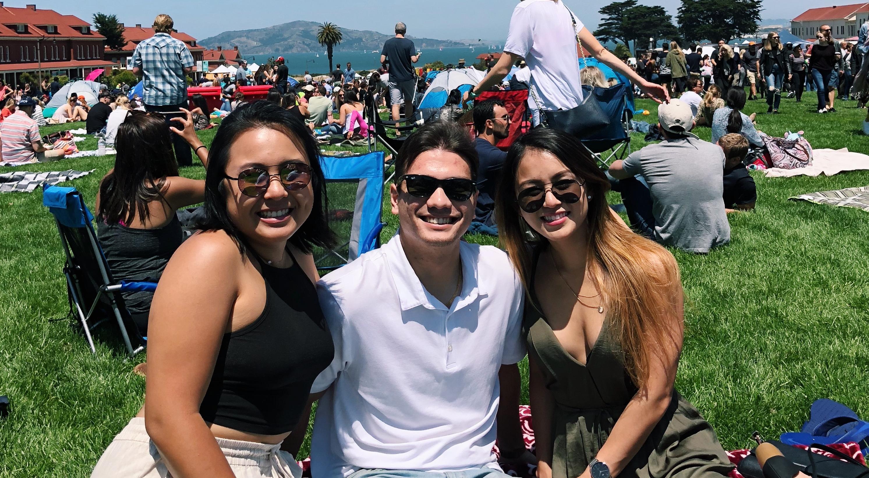 Three people smiling at a picnic in a park on a sunny day.