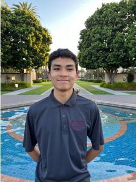 A young man standing outside near a pool, trees in the background.