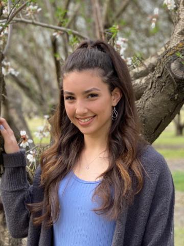 Person smiling outdoor with long, wavy hair and trees in the background.