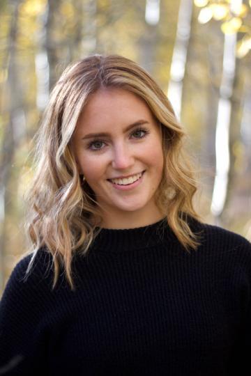 A woman smiles, standing in front of trees.