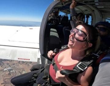 Person wearing skydiving gear, smiling, sitting in the open door of an airplane.