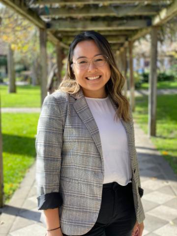 A woman wearing a blazer stands outdoors, smiling.