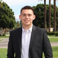 A person in formal attire standing outside with palm trees in the background.