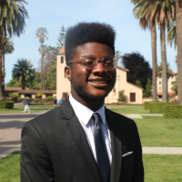 A person in a suit standing outdoors with palm trees in the background.
