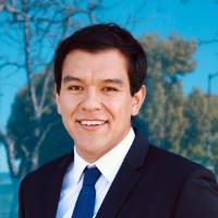 A man in a suit smiling outdoors with a blurred background.