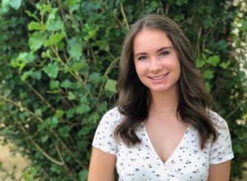 A woman in a white dress standing in front of greenery.