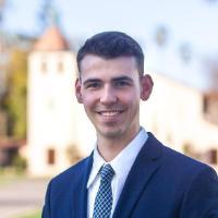 A person in a suit standing outside with buildings and trees in the background.