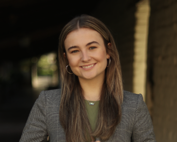 A person smiling outdoors, wearing a gray blazer.