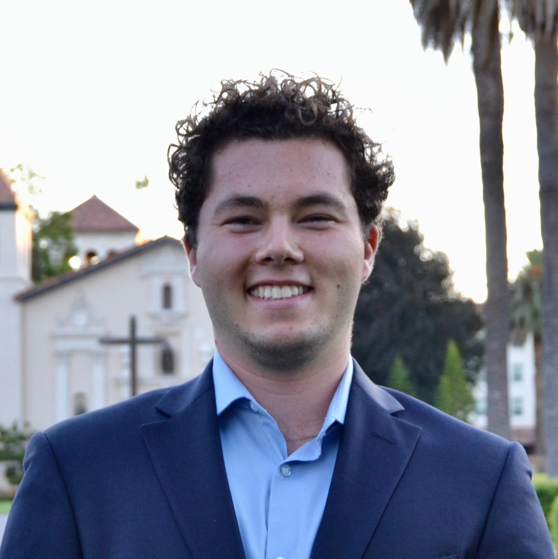 A person in a suit standing outdoors with houses and palm trees behind.