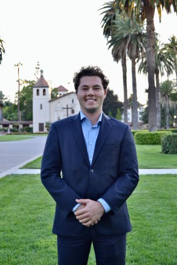 A person in a suit standing outdoors with houses and palm trees behind.