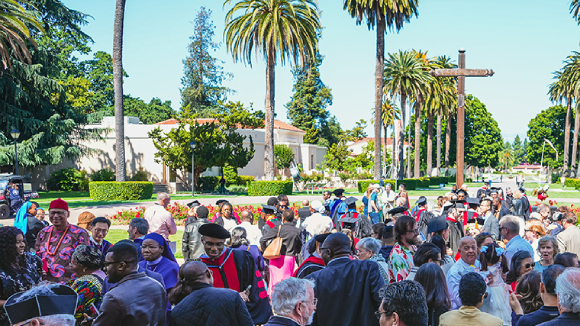 A group of people, some in religious garments and some in graduation regalia, congregate in front of a large wooden cross.