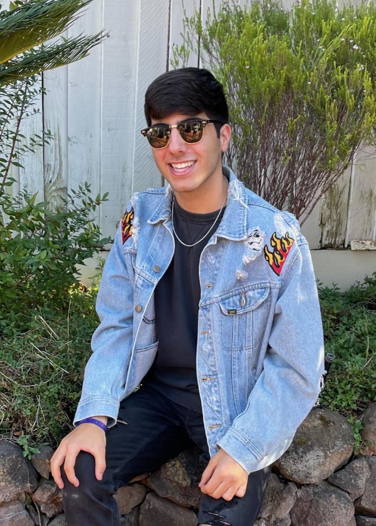 Person in blue denim jacket and sunglasses sitting outdoors near green plants.