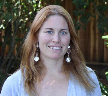 A person smiling, wearing a gray top and large earrings, outdoors.