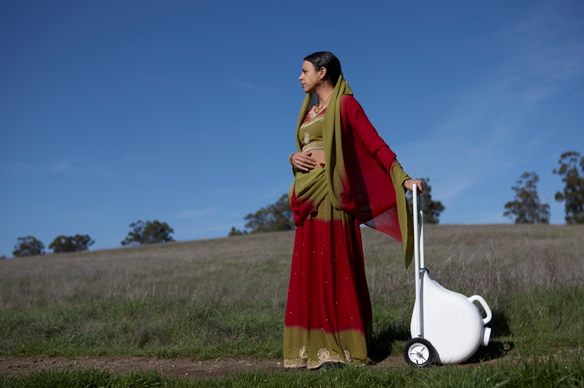 A woman in traditional attire stands outdoors holding a water jug.