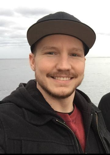 A person smiling outdoors wearing a dark jacket and a cap.