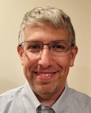 A person smiles at the camera, wearing glasses and a striped shirt with a tie.