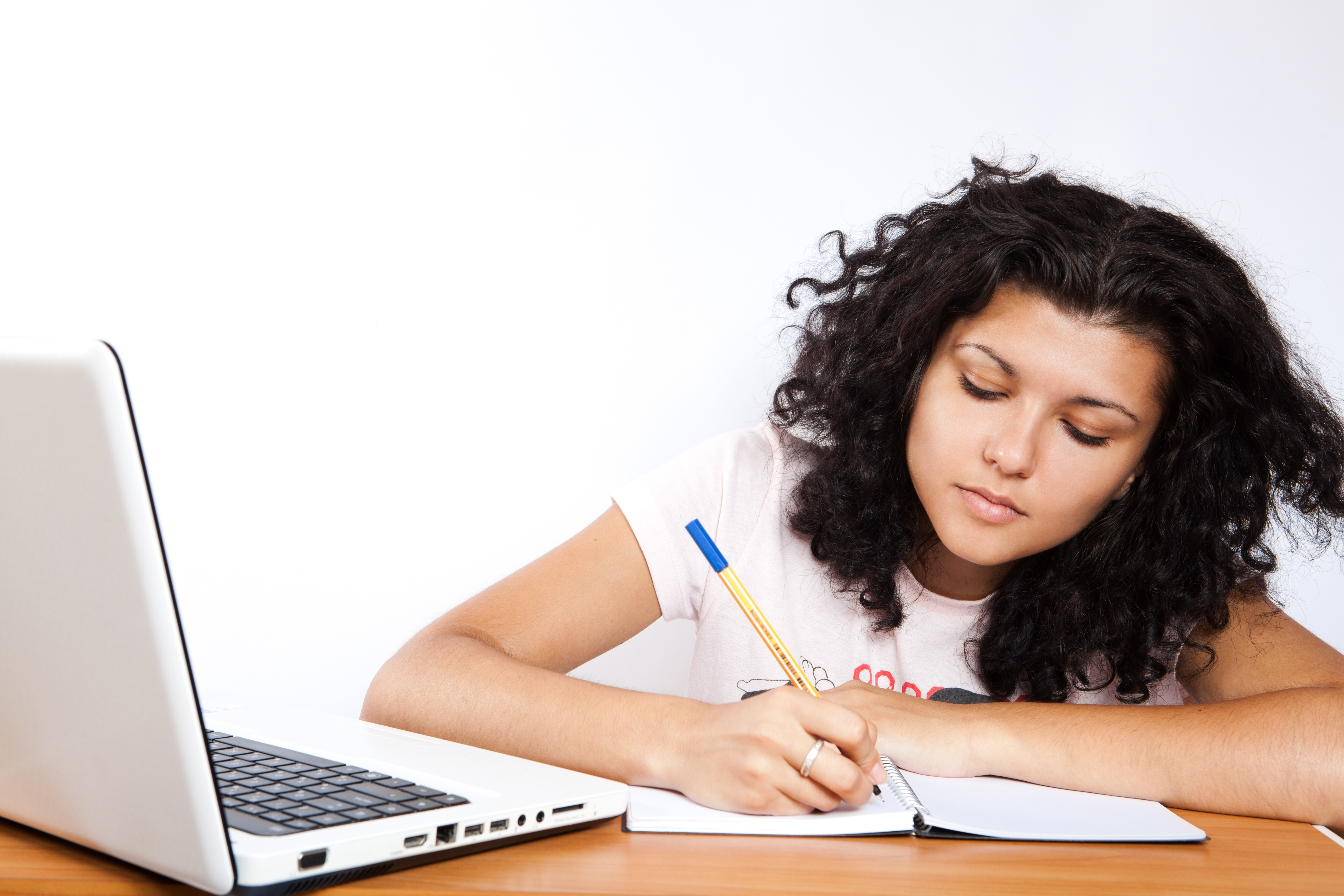 A student writes in a notebook beside a laptop.