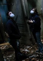 Two people wearing masks in a dark, possibly abandoned, area.