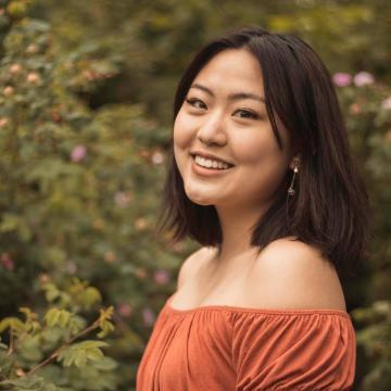 A woman in an orange top smiling in front of greenery.