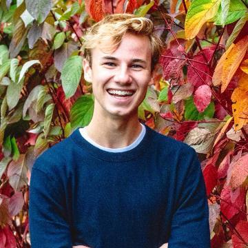 A smiling person stands in front of colorful autumn leaves.