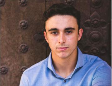 A person wearing glasses and a blue shirt in front of a wooden wall.