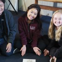 Three people smiling and sitting cross-legged, showing a piece of paper with a drawing.