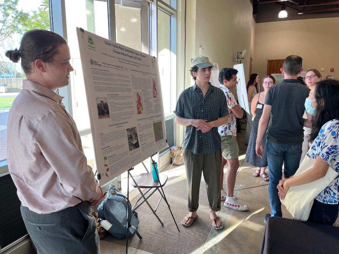 two students on either side of a poster on an easel are speaking about their research