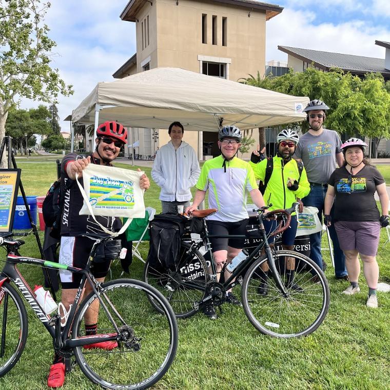 A group of people wearing bike helmets and standing with their bikes in front of a table with a canopy overhead