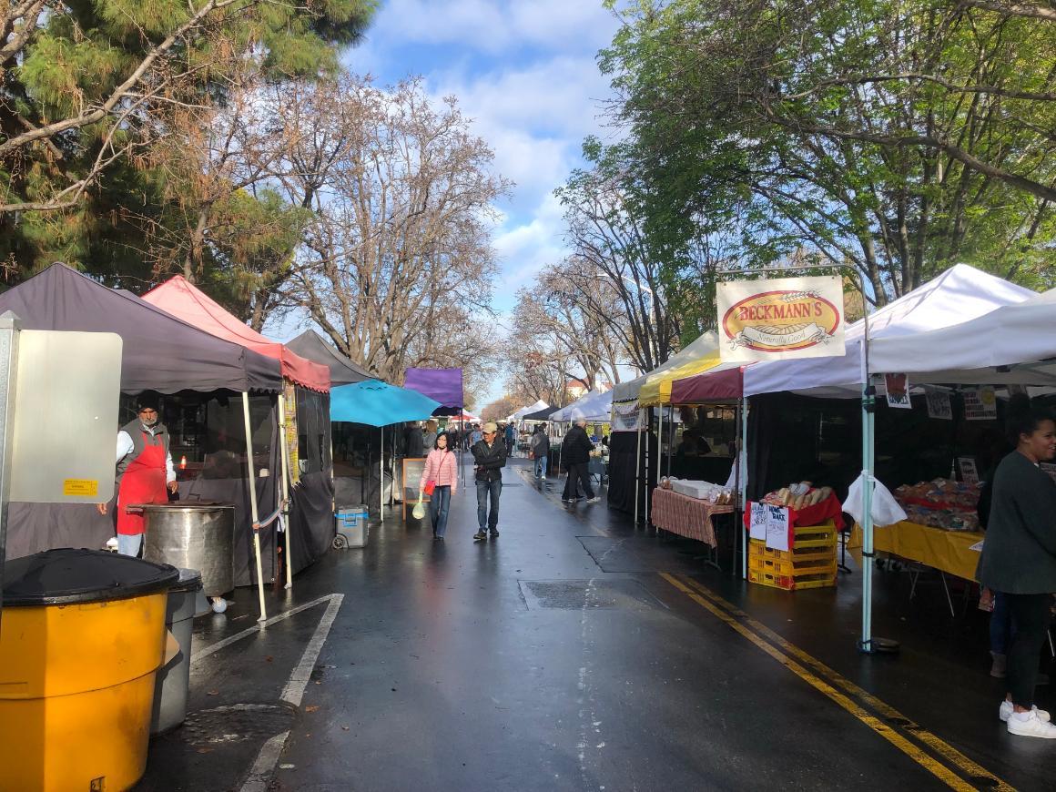 Outdoor farmers market with people walking and vendor stalls on both sides of the pathway.