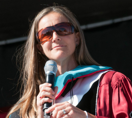 Person wearing academic regalia speaking into a microphone outdoors. image link to story