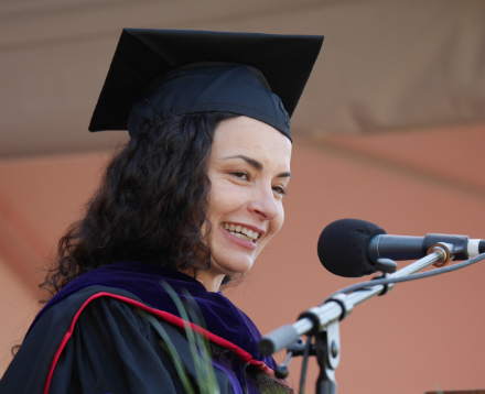 A person in graduation attire speaks into a microphone. image link to story