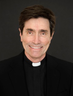 A person wearing a clerical collar and black attire, smiling in front of a dark background.