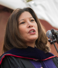 A person speaking, wearing academic regalia, with a microphone in the background. image link to story