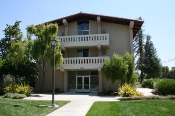 A three-story building with balconies, surrounded by grass and trees.