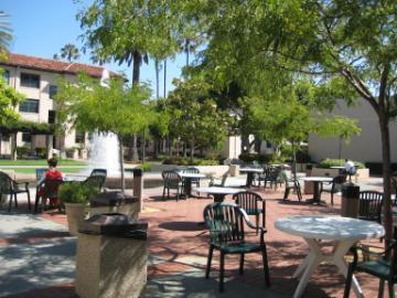 Outdoor terrace with tables, chairs, trees, and people under a clear sky.