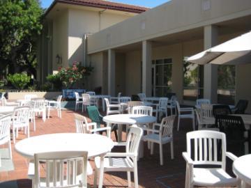 Outdoor patio with white tables and chairs under a covered area on a sunny day.
