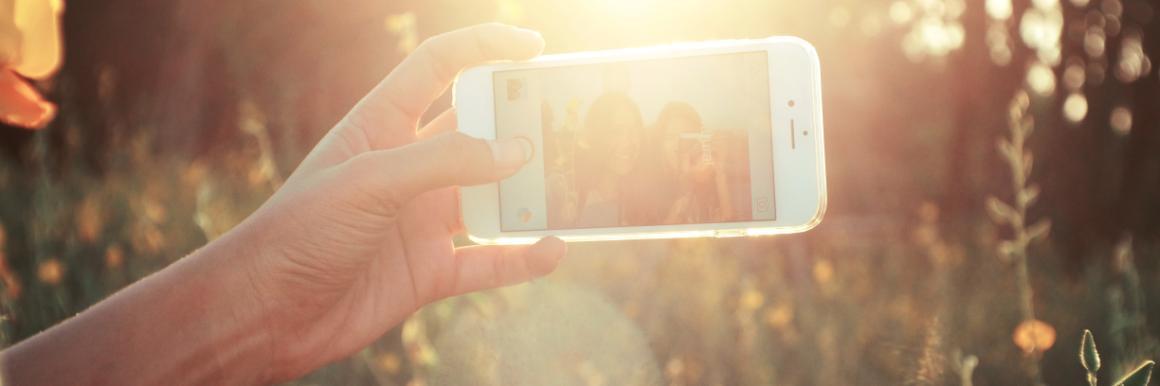 A hand holding a smartphone capturing sunlight through trees.