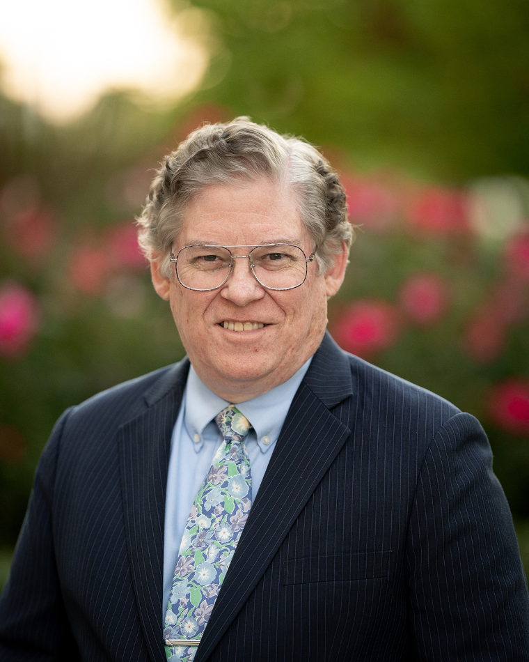 A man in a suit and glasses standing in a rose garden.