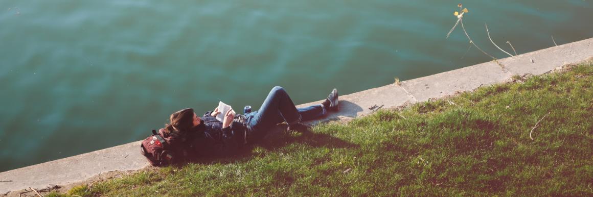 Young adult woman laying by the water reading a book