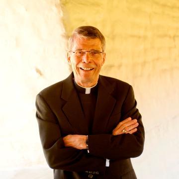 A priest in a black suit smiling with arms crossed.