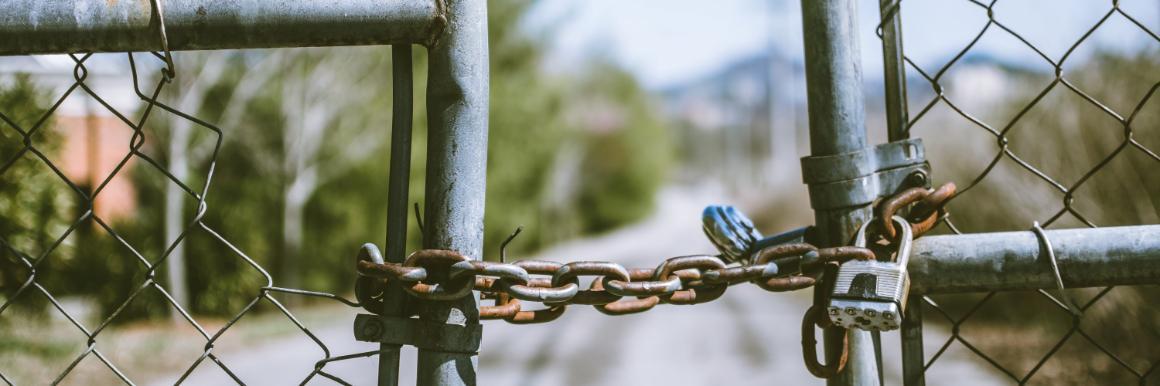 A gate with a chain holding the two doors together by a padlock in front of a blurred road and outdoor background