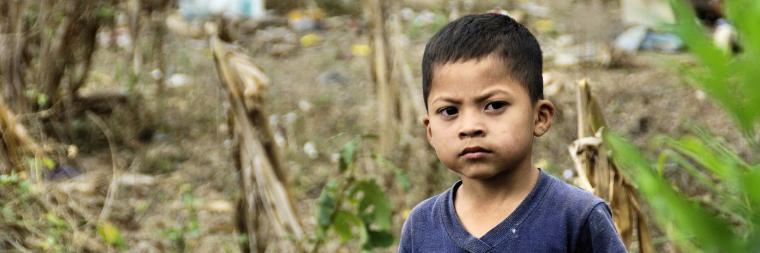 A young boy stands outdoors with trees and soil in the background.