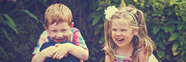 Two children smiling and sitting side-by-side outdoors.