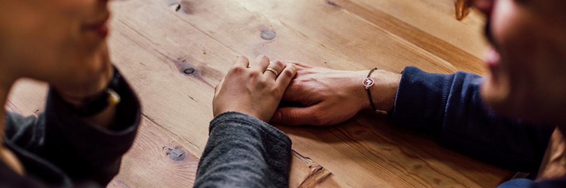 A man and a woman sitting at a table blurred in the forefront, her hand comfortingly on top of his