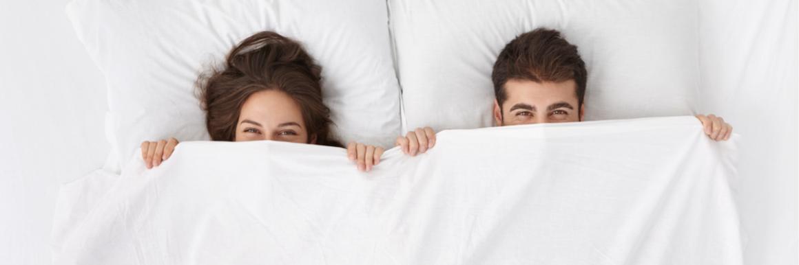 A happy couple are in bed, peeping over the covers.  